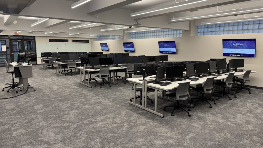 Rows of computer lab tables with a main lectern and displays at the end.
